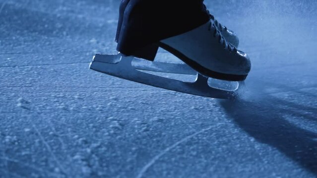 Detail shot of women's legs in figure skating skates sliding on cold ice in arena. A professional sportswoman trains in the dark with blue light. Woman slides on ice and stops. Close up. Slow motion.