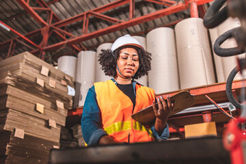 Fototapeta na wymiar Engineer asian and african woman wearing safety helmet and vest holding clipboard and take note on the paper in the automotive part warehouse.Products and corrugated cardboard. 