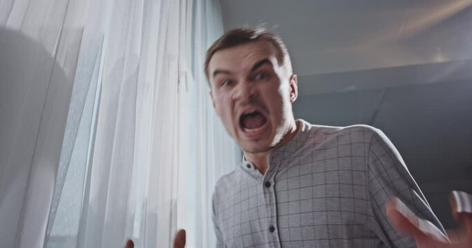 Angry man looking out window. From below mad male in checkered gray shirt looking at window with curtains then screaming in camera in anger at home in daytime
