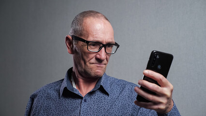 Senior man pensioner with bad eyesight in elegant black-rimmed glasses tries to read message in modern smartphone on grey background closeup.