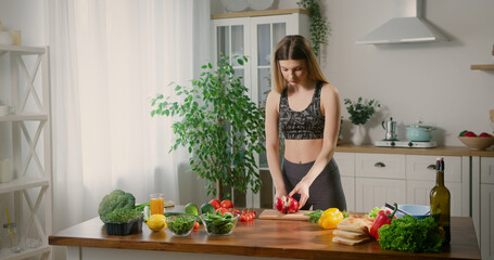 Fit young woman cooking healthy food at home. Slim attractive lady cutting crispy bell pepper, making fresh salad. Various vegetables on wooden table.