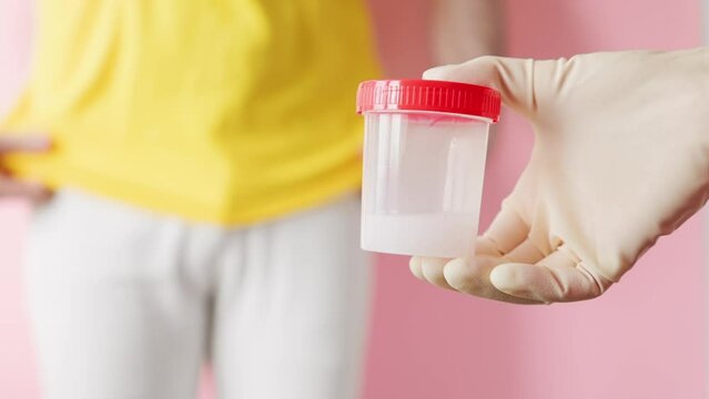 Doctor's hand in medical gloves is a close-up, holding a plastic jar with a sample of sperm. In the background, an unfocused man is buttoning his pants. The concept of sperm donation and analysis