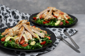 Chicken salads with avocado, rucola and cherry tomatoes in a black plate on gray stone background.