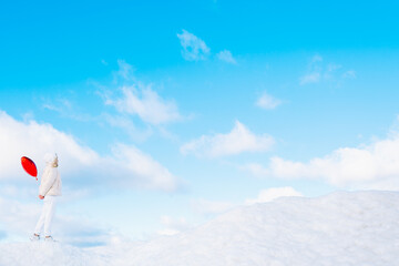 Portrait of young woman in white clothes with red heart balloon walking on snow cover with blue cloudy sky on background. Sky merges with earth near snowdrifts. Side view. Copy space. Minimalism