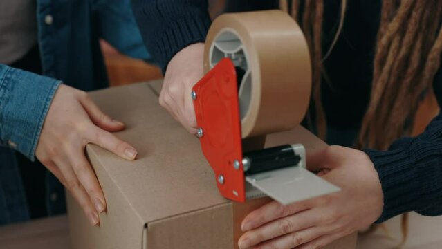 Cropped view of the young female warehouse workers seals parcels with tape package, while preparing delivery parcel box at the home office. Post service and small business concept.