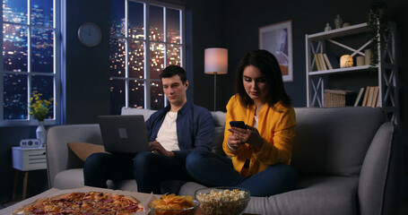 Two young people spending evening time at home, using gadgets for work or personal needs. Married couple resting on the couch, sitting silently in living room. Man with laptop. Woman with phone.