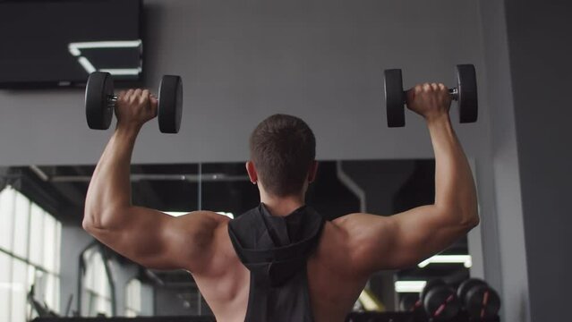 Young male bodybuilder trains in gym, lifting two dumbbells with two hands, back view. Male athlete is engaged in training at gym with dumbbells lifting them