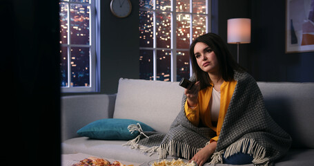 Extremely bored young woman student watching tv switching channels with remote control. Depressed unmotivated lady wrapped in soft rug sitting on couch at home.