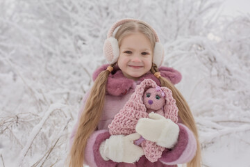 a blonde girl in a pink fur coat plays in a snowy winter forest with a plush toy