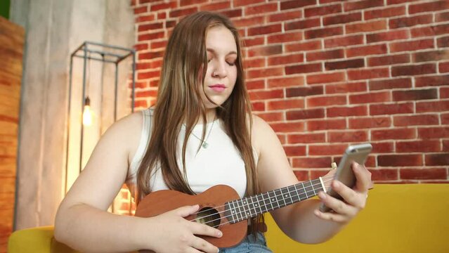 Young woman with ukulele watches training video on her phone, puts phone down and begins practice, front view. Concept of self learning
