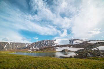 Colorful green landscape with mountain lake in sunlit meadow against high snowy mountain range under cloudy sky in changeable weather. Vivid sunny scenery with snow mountains reflected in alpine lake.