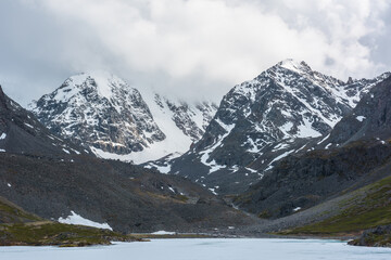 Dramatic mountain landscape with frozen alpine lake and high snow mountains and sunlit big rocks among low clouds. Atmospheric scenery with ice lake on background of large snowy mountains in overcast.