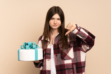 Young Ukrainian girl holding a big cake isolated on beige background showing thumb down with...