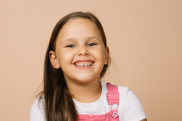 Little girl with bright shining eyes and excited blissful smile with teeth looking at camera wearing bright, pink jumpsuit and white t-shirt on beige background.