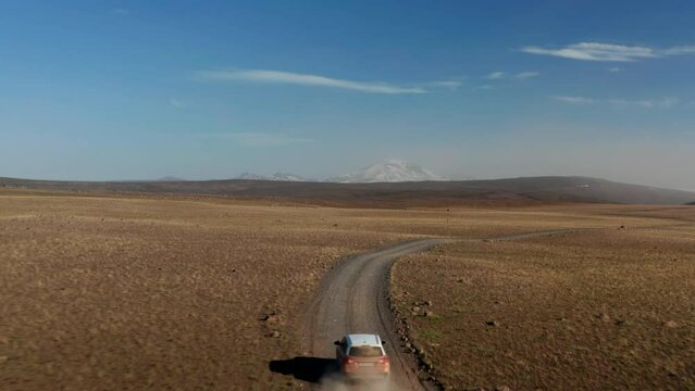Fly above plains, revealing car driving on dirt road. Snow capper mountain peaks in distance. Iceland