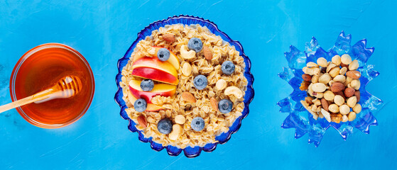 Fototapeta na wymiar Oatmeal porridge with apple slices, berries and nuts. Bowl of oatmeal and honey on a blue background. Healthy breakfast concept. Top view