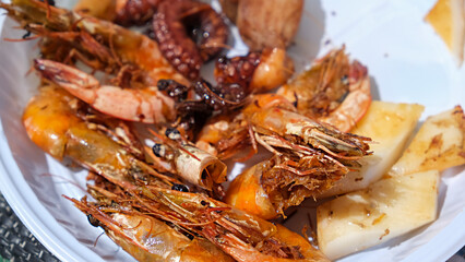Disposable plate with a mix of grilled seafood shrimp, octopus, lobster, squid. BBQ exotic food on the beach.