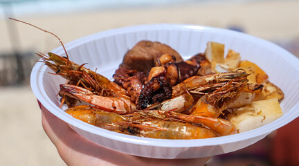 Disposable plate with a mix of grilled seafood shrimp, octopus, lobster, squid. BBQ exotic food on the beach.