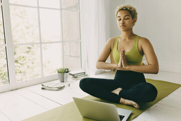 Dark-skinned blonde curly-haired female sitting at yoga mat in prayer position and eyes closed, meditating, following online instructor, sitting in front of opened laptop near panoramic window