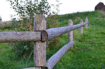 A fence made of wooden logs, against the background of a trail. In the background is a haystack. Village.