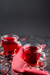 Hibiscus tea in glass cup and burgundy napkin. Cup of red hibiscus tea and dry hibiscus petals on dark background. Healthy slimming drink. Copy space