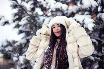 Beautiful young brunette girl with long hair in a white fur coat and a white hat has fun at the resort. Portrait outdoor photography