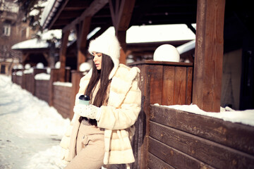 Beautiful young brunette girl with long hair in a white fur coat and a white hat has fun at the resort. Portrait outdoor photography
