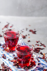 Obraz na płótnie Canvas Hibiscus tea and dry hibiscus petals on gray background. Two glass cups of red hibiscus tea on blue napkin. Top view. Copy space