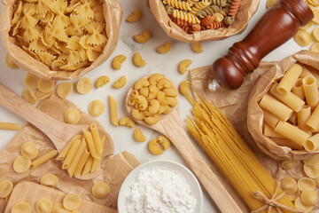 pasta concept some types of pasta being organized on wooden spoons and in paper bags while some and other ingredients being put on white background