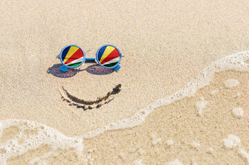 Fototapeta na wymiar A painted smile on the sand and sunglasses with the flag of Seychelles. The concept of a positive and successful holiday in the resort of Seychelles.