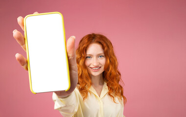 Portrait of excited redhead girl holding big smartphone with white blank screen in hand, showing...