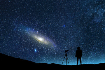 The silhouette of a man with a camera stands on a hill in the starry night sky. The bright Andromeda galaxy and meteors behind it.