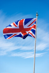 Union Jack Flag. Close-up of a national UK flag with flagpole, blowing in the wind against a blue sky with clouds and copy space.