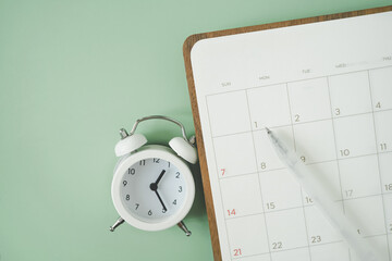 white pen on blank white planner and analog alarm clock on green background, top view