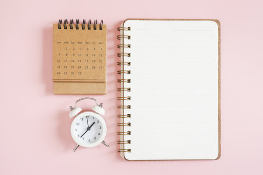 top view of blank opened note book, grunge brown monthly calendar and analog alarm clock on pink background