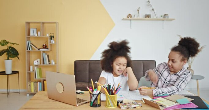 Smiling mixed race junior schoolgirls with kinky hair draw on paper with colorful pencils near laptop at table in children room