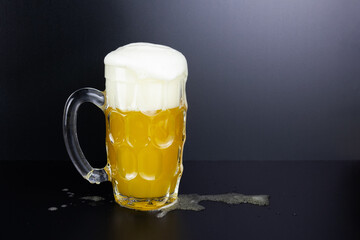 Glass mug with beer and white foam on black background, some spilled drink