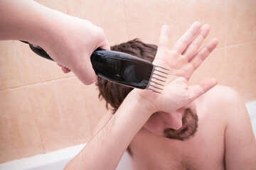 Shaggy man refusing shaving, hiding his face behind palm from electric razor in female hand,...