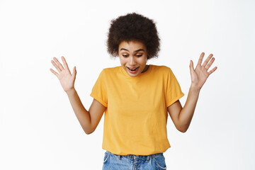 Portrait of surprised african woman raising hands up, looking down in awe and amazement, seeing something exciting below, scream with enthusiastic face, white background