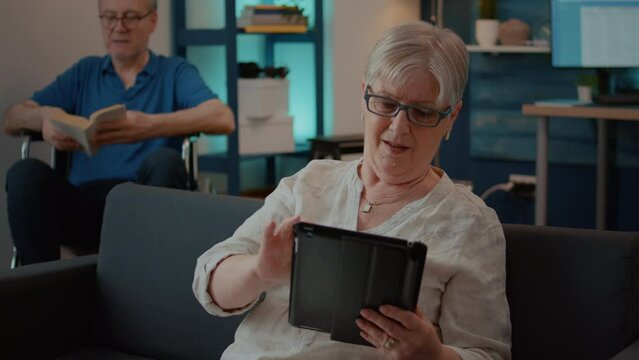 Retired woman using digital tablet to enjoy free time at home, browsing internet. Old person with crutches enjoying retirement with device on couch in living room, relaxing for recovery.