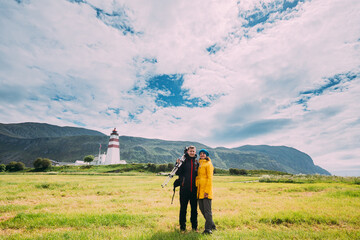 Alnesgard, Godoya, Norway. Man And Woman Young Adult Caucasian Tourists Travelers Couple Posing Against Old Alnes Lighthouse In Summer Day In Godoy Island Near Alesund Town. Alnes Fyr. - obrazy, fototapety, plakaty