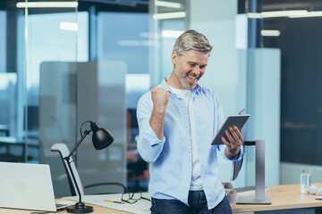 Senior and experienced business man uses a tablet to work, gesture of victory hand, received good...