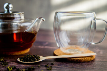 A wooden spoon with tea lies against a dark wood background, a mug and a teapot are visible from behind, a photo in dark colors. High quality photo