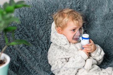 Cute baby boy makes inhalation with a nebulizer equipment. Sick child holding inhalator in hand and breathes through an inhaler at home - obrazy, fototapety, plakaty