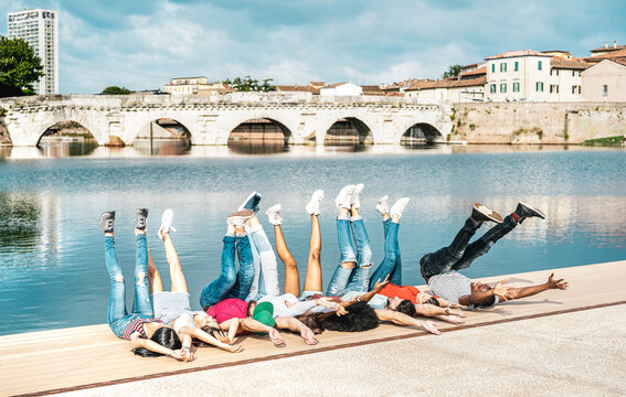 Multiracial milenial friends cheering at city docks lying on the floor - Happy guys and girls having fun around old town place - University students on travel life style vacation - Bright vivid filter