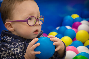 Fototapeta na wymiar A little boy in a blue sweater with glasses plays with multi-colored plastic balls in the playroom.