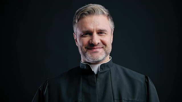 Handsome priest smiling on camera, friendly to parishioners, black background