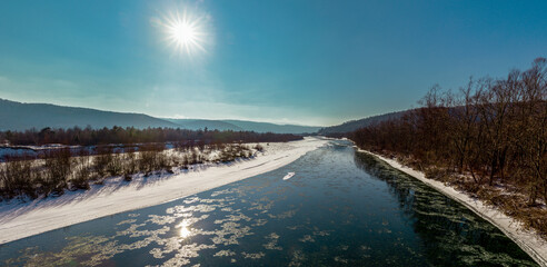 Winter frosty day - panorama. The sun in the blue sky over the mountains and the mountain river on a frosty winter day. Winter panorama of the mountain river Stryi, Carpathians, Ukraine. 