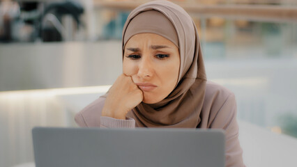 Young arab sorrowful woman in hijab reading message on laptop gets bad news denied bank loan job...