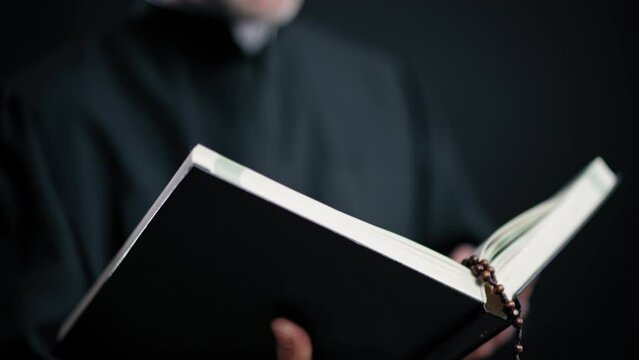 Serious Catholic priest reading Bible against black background religious service